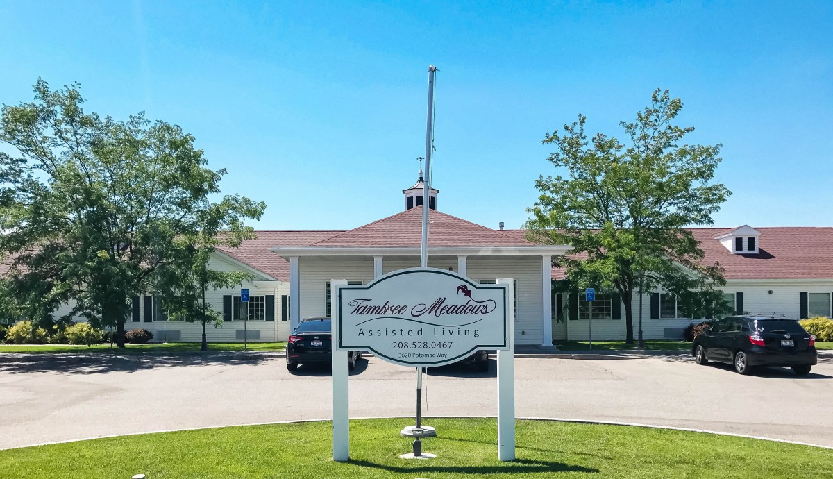 A building with a sign labeled 'Woodside Meadows' in front, surrounded by trees and under a clear blue sky.