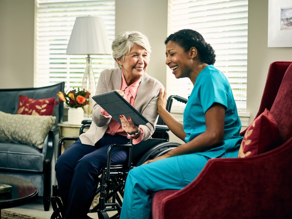 A nurse interacts with a senior woman in a wheelchair, both looking at a tablet in a cozy home setting.