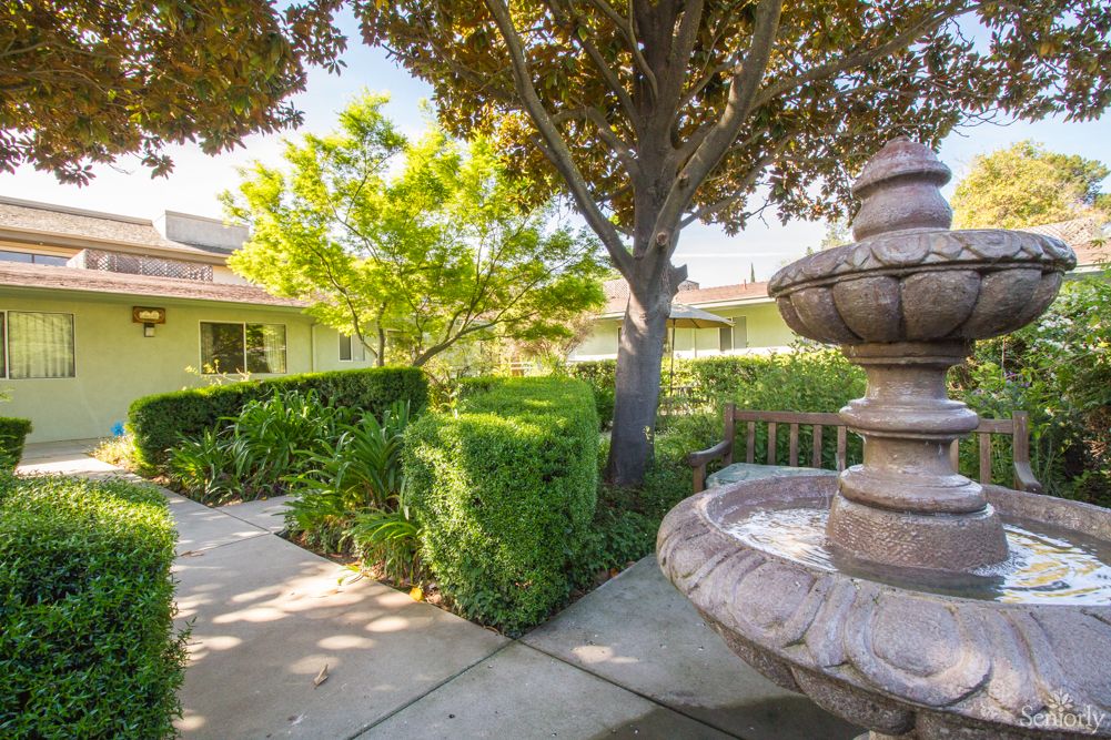 A serene outdoor garden with a stone fountain, surrounded by lush greenery and hedges under the sunlight.