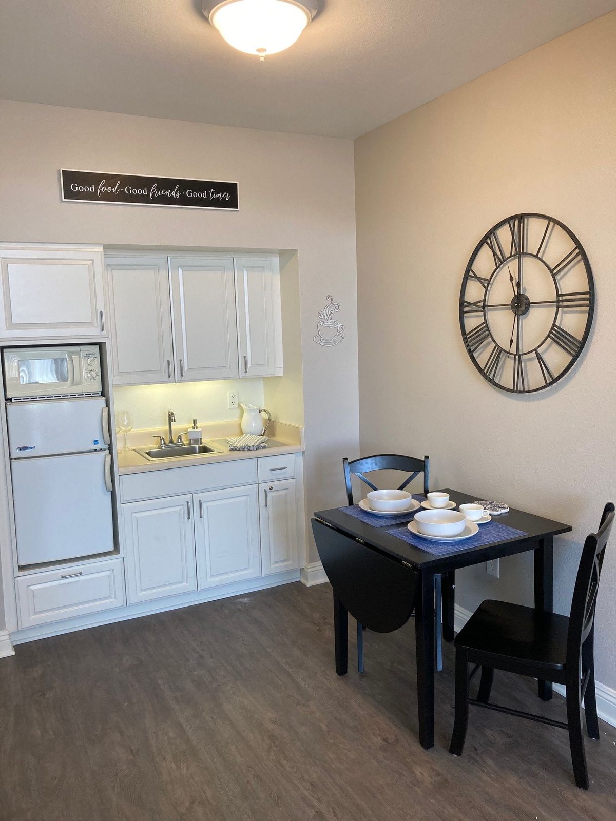 A compact kitchen space with white cabinets, a small dining table set for two, and a large wall clock.