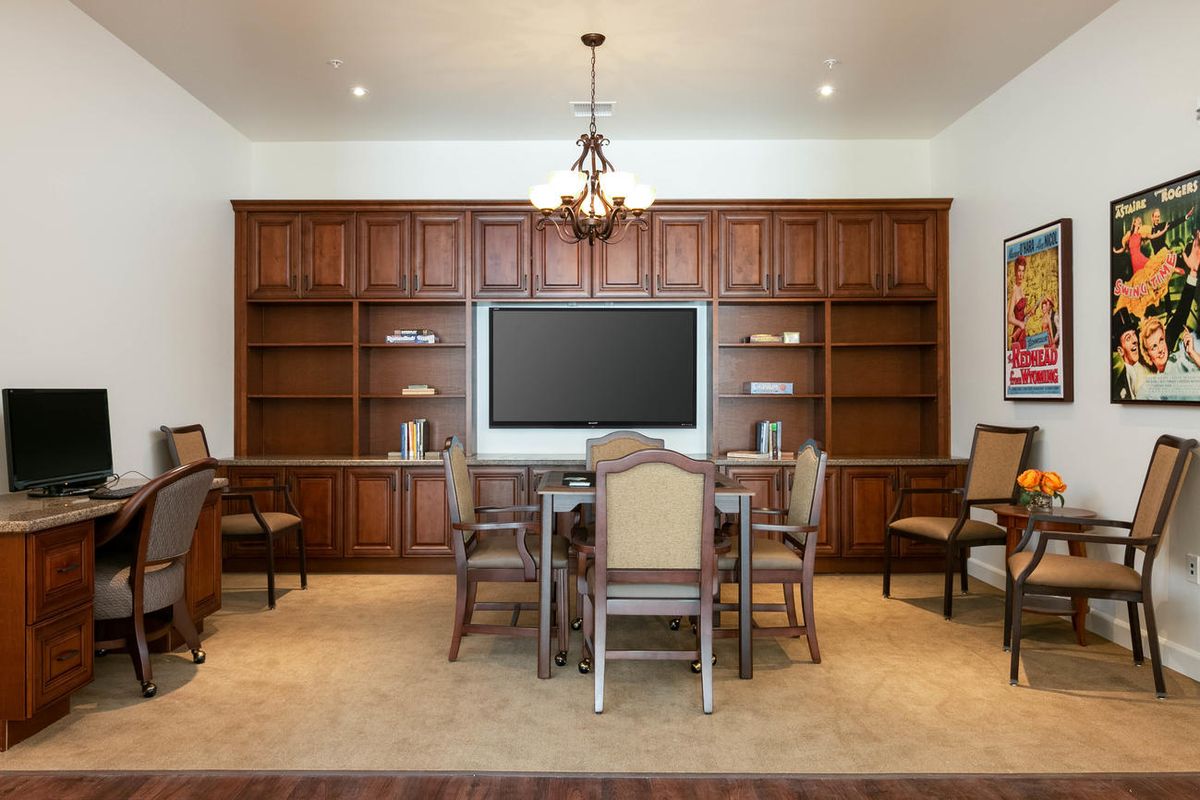 A dining room with wooden cabinets, a central table and chairs, a chandelier, and a large television.
