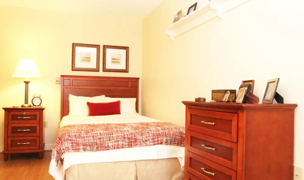 A cozy bedroom with a neatly made bed, a wooden dresser, and decorative items on a shelf.
