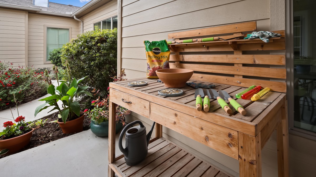 A wooden potting bench is set up with gardening tools and a watering can, surrounded by potted plants.