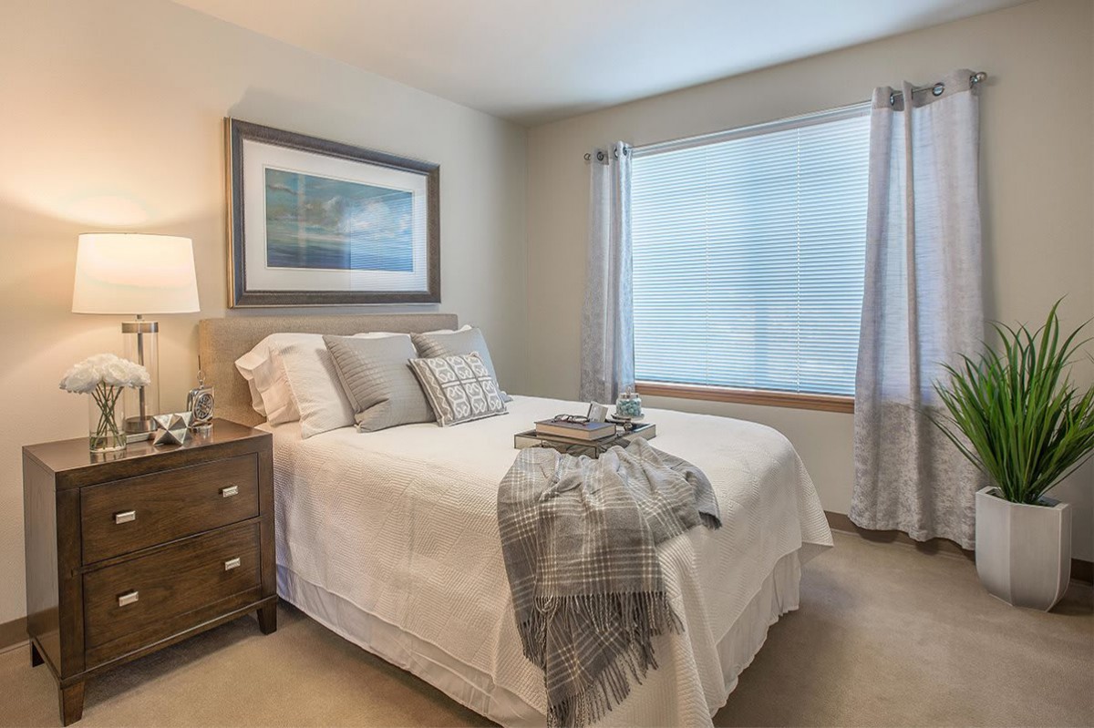 A cozy and elegantly decorated bedroom with a neatly made bed, wooden nightstand, and large window for natural light.