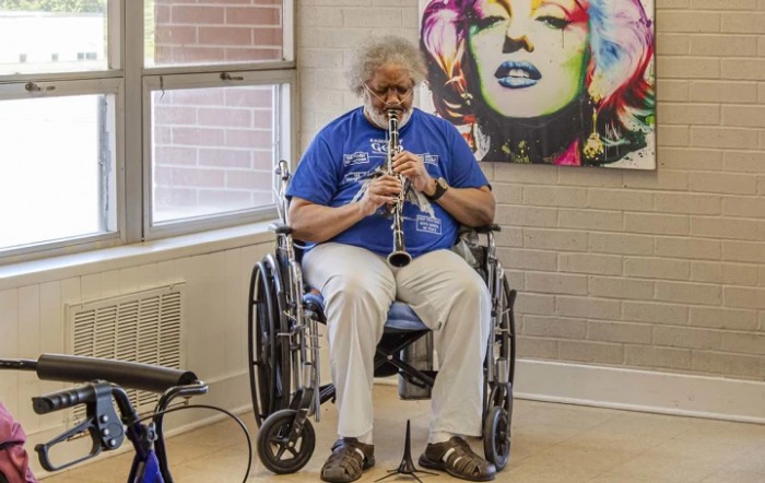 A person in a wheelchair plays the clarinet indoors beside colorful wall art.