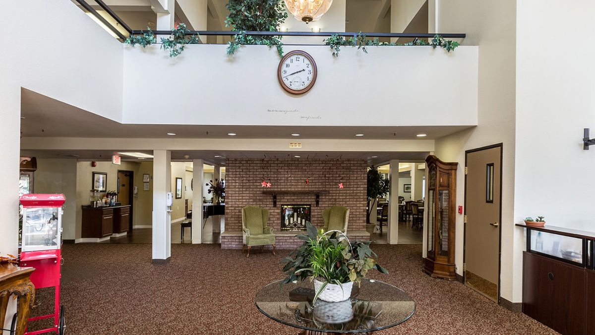 A hotel lobby featuring a central table with plants, a fireplace with seating, and a wall clock above.
