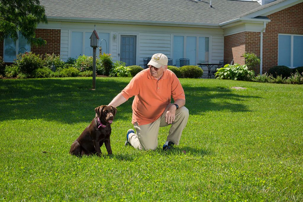 A man kneels on a green lawn, affectionately petting a brown dog in front of a house.