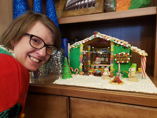 A cheerful individual poses with a beautifully crafted gingerbread house, showcasing festive spirit.