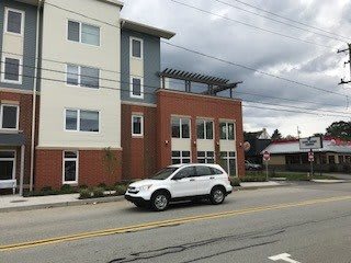A white car is parked on a street in front of a modern multi-story building under an overcast sky.