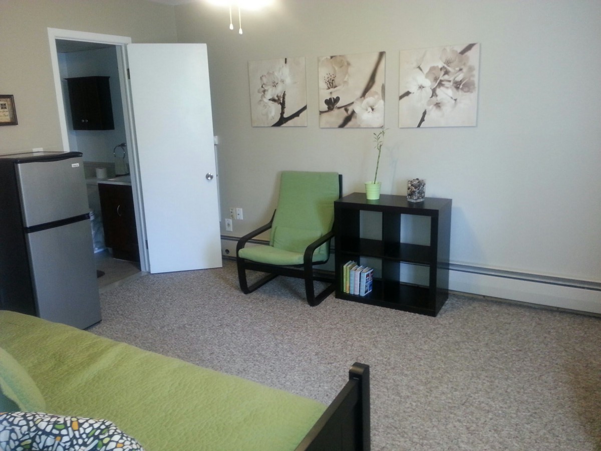 A tidy bedroom featuring a green chair, a black shelf with decor items, and floral artwork on the wall.