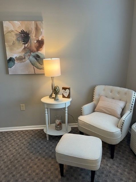 A cozy living room corner featuring a comfortable armchair, a side table with a lamp, and decorative artwork on the wall.
