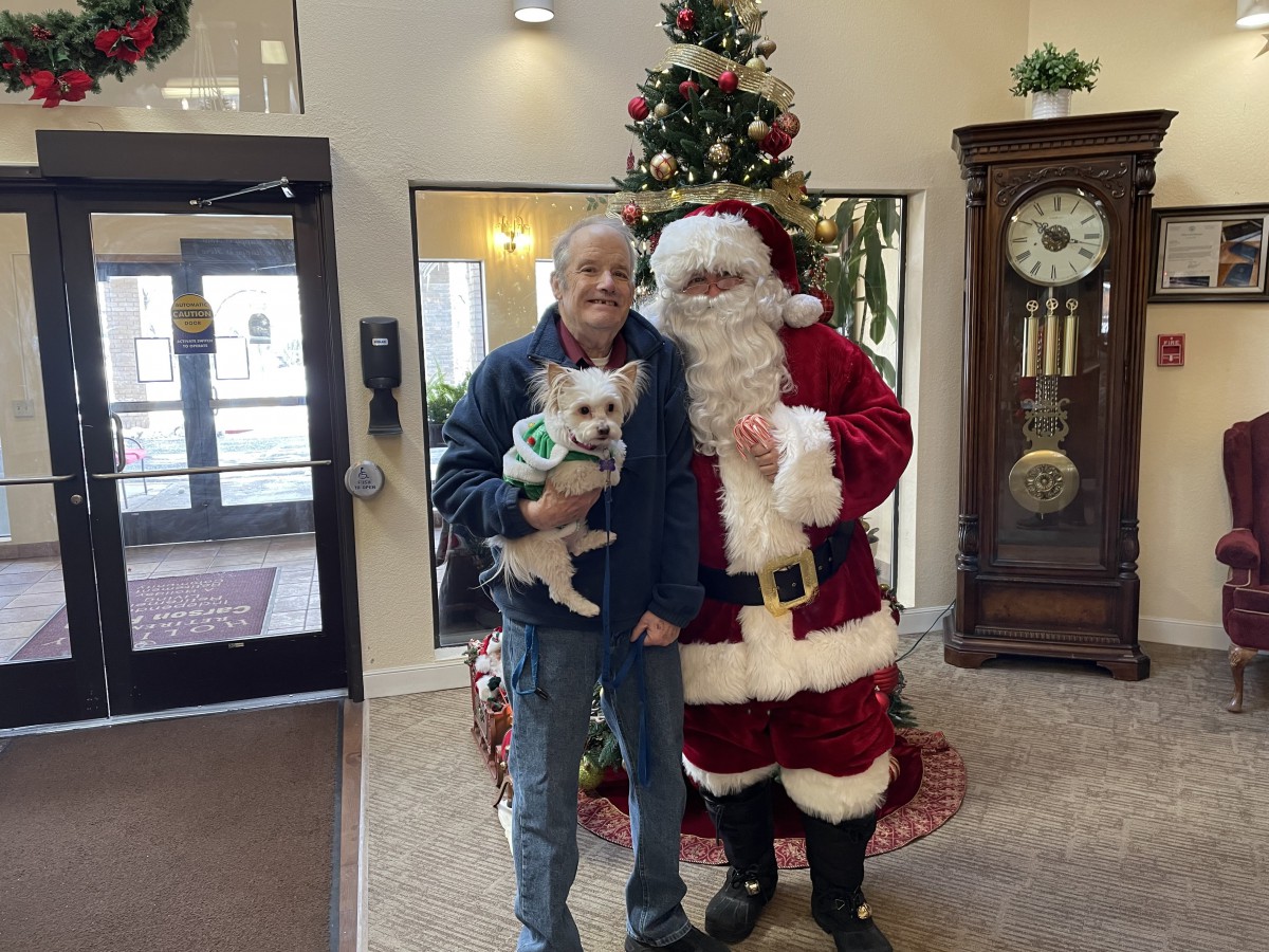 A person holding a dog stands next to Santa Claus in a festive indoor setting decorated for Christmas.