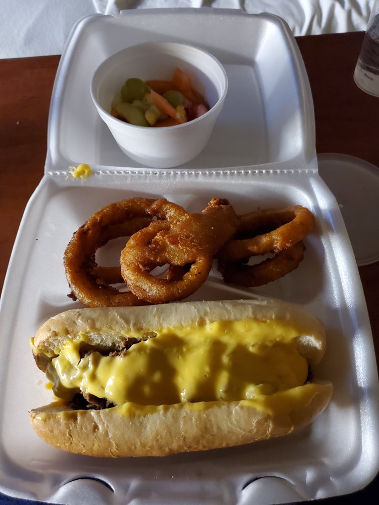 A meal consisting of a cheese-covered hot dog, onion rings, and a side dish served in a disposable container.