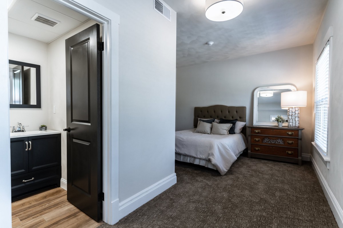 A modern bedroom with a carpeted floor, featuring a bed and a dresser with a mirror.