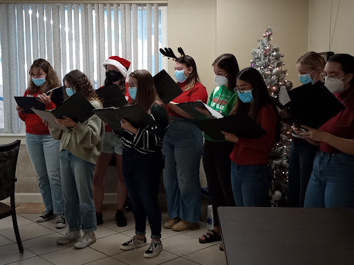 A choir performs festive songs in holiday attire, surrounded by a decorated Christmas tree.