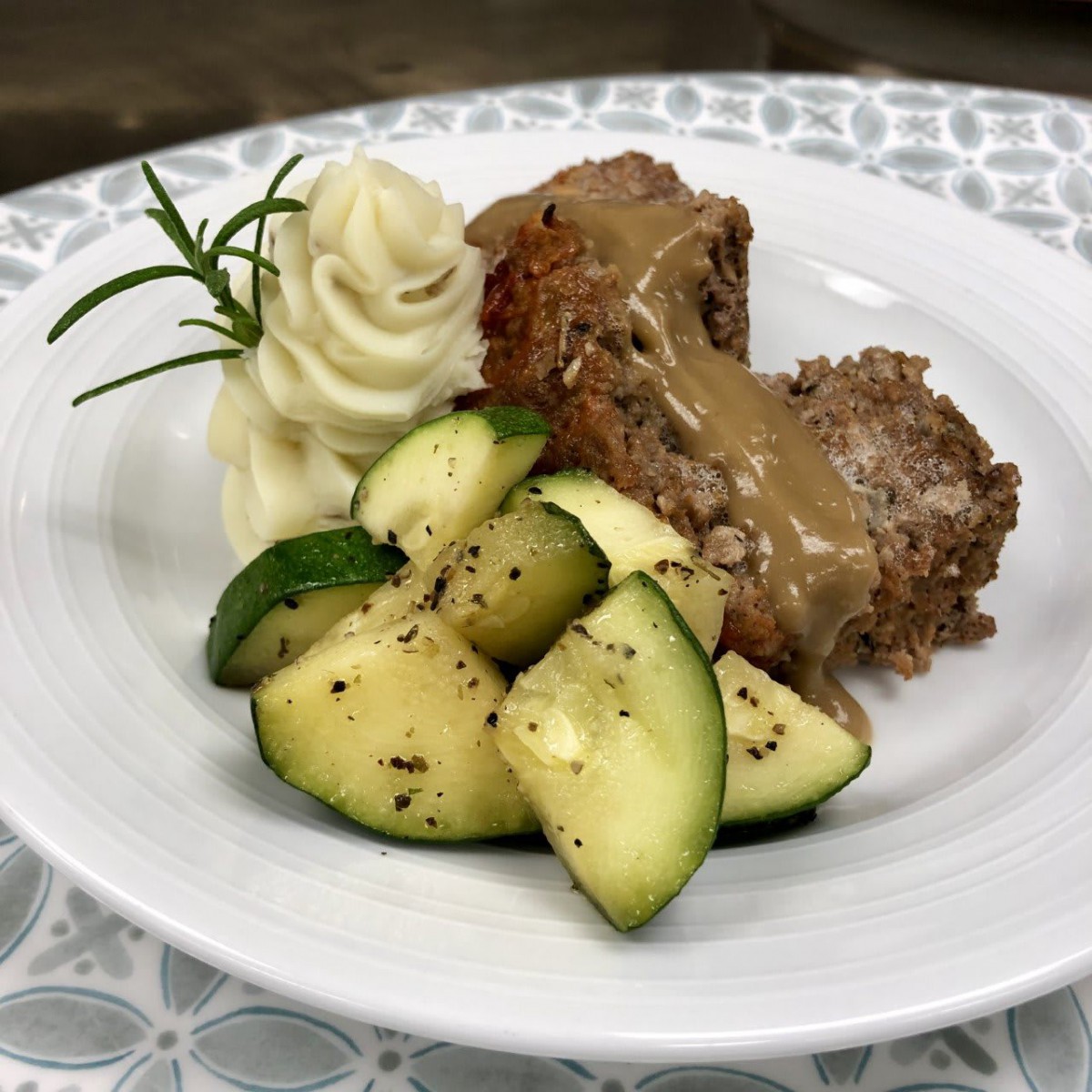 A beautifully plated dinner featuring meatloaf with gravy, creamy mashed potatoes, and seasoned zucchini slices, garnished with a sprig of rosemary.