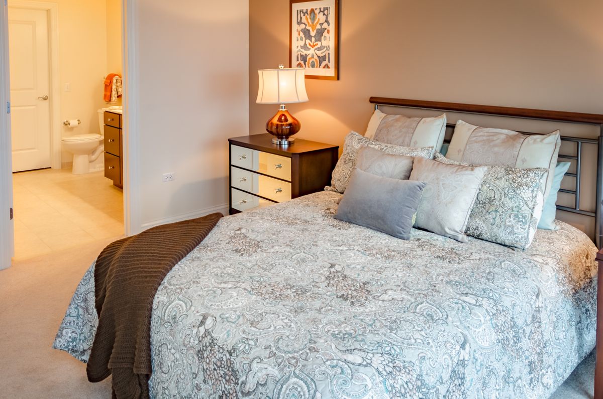 A cozy bedroom with a neatly made bed featuring multiple pillows, a patterned comforter, and a nightstand with a lamp.