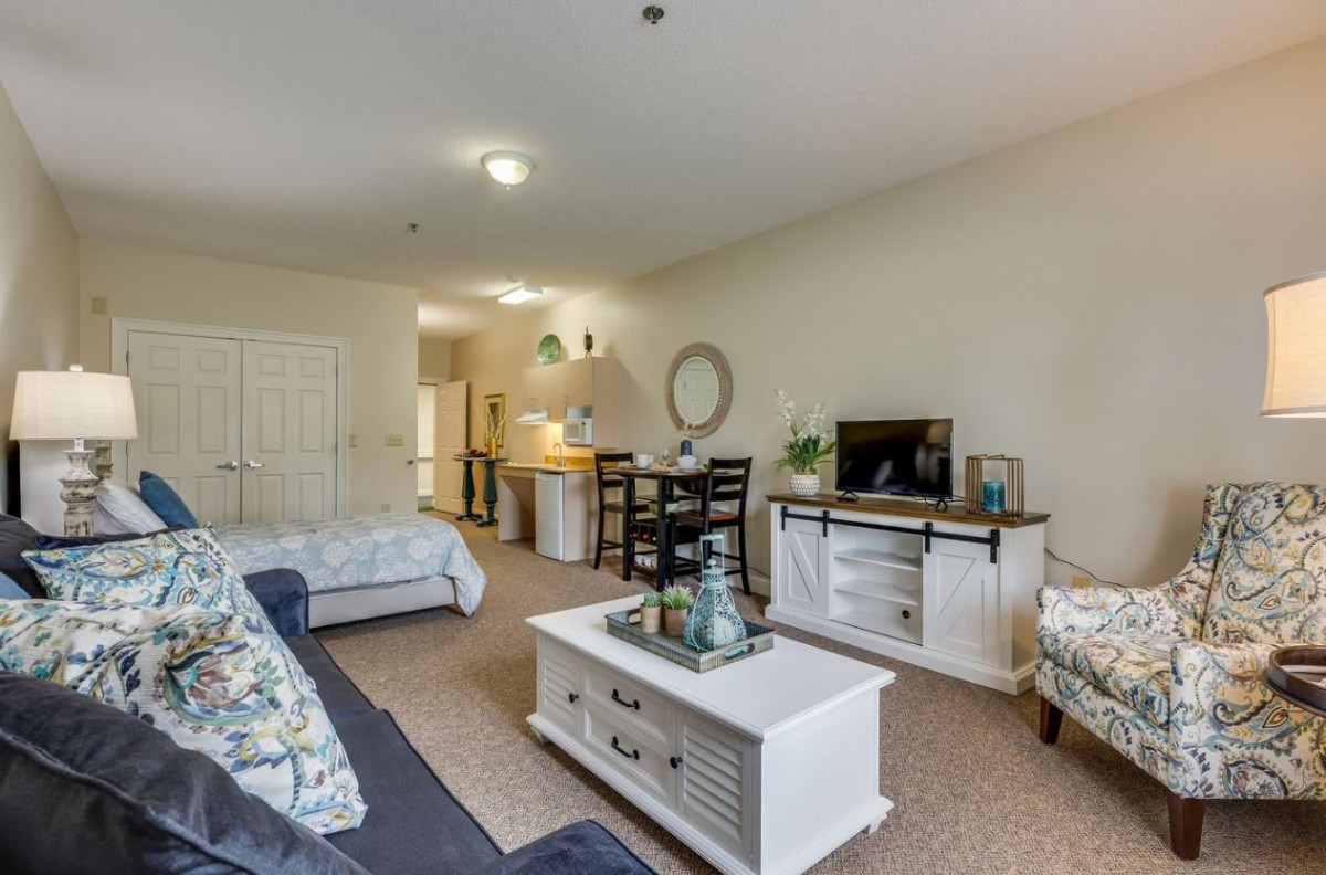A cozy apartment living area featuring comfortable seating, a TV unit, and an open kitchen in the background.
