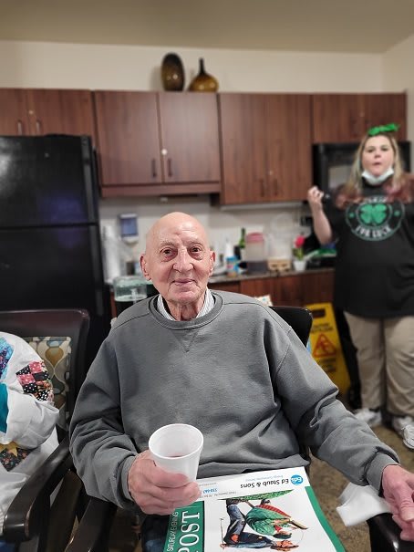 An elderly man smiling while holding a cup, seated in a kitchen setting with a caregiver standing nearby.
