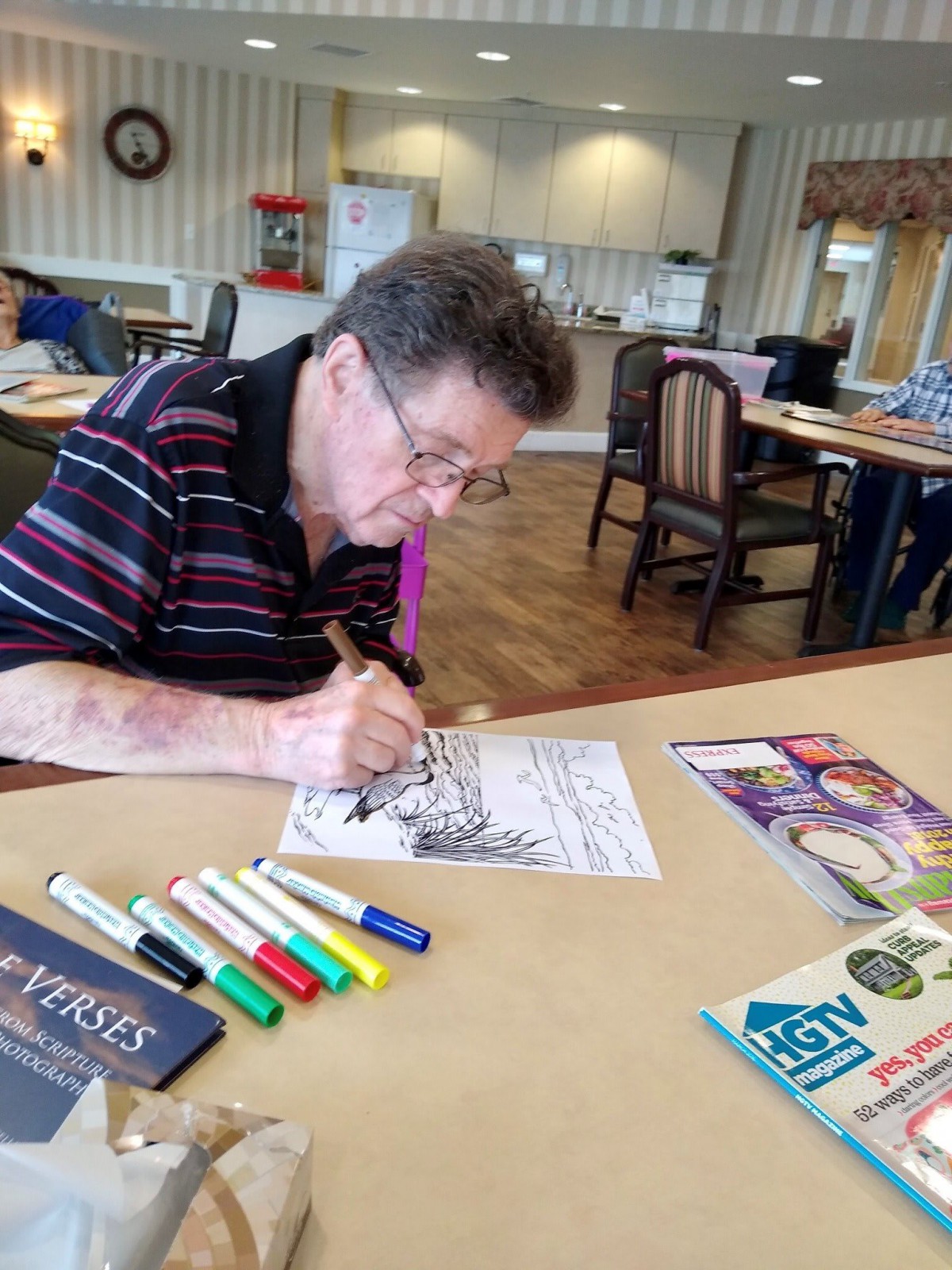 An elderly man focuses on coloring a picture with markers, engaging in an artistic activity indoors.