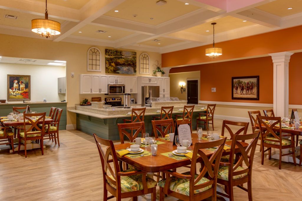 A warmly lit restaurant dining area featuring neatly arranged tables and chairs, with an open kitchen in the background.