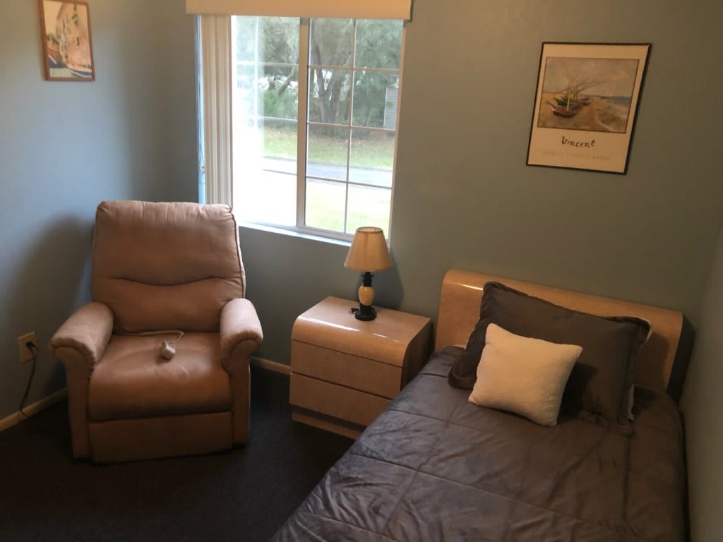 A cozy bedroom with a bed, nightstand, and reclining chair next to a window.