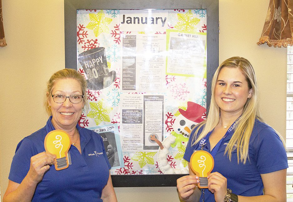 Two women smiling while holding awards in front of a decorated bulletin board.