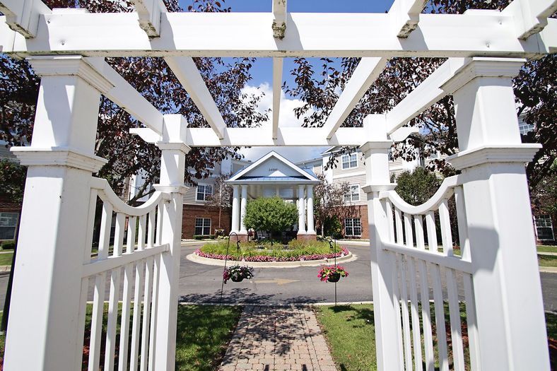 A scenic view of a white pergola leading to a building surrounded by trees and landscaping.