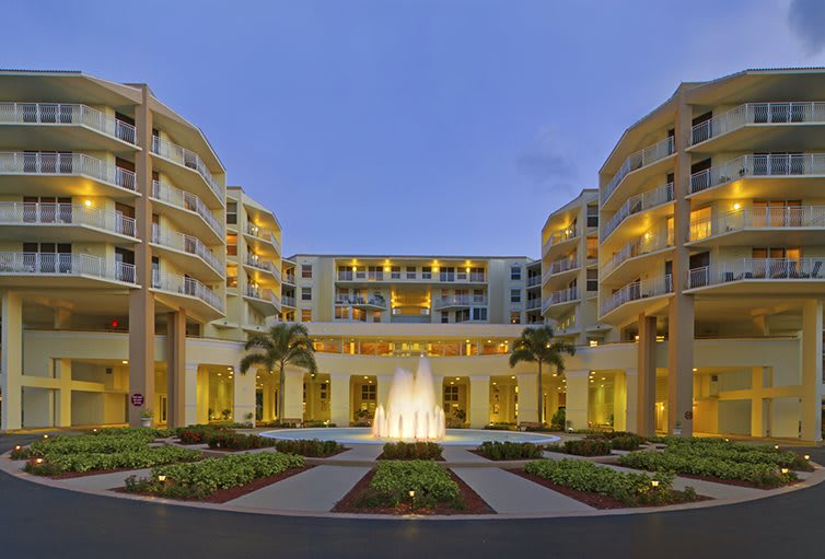 Luxurious hotel exterior with a central fountain, surrounded by landscaped gardens and palm trees in the evening.