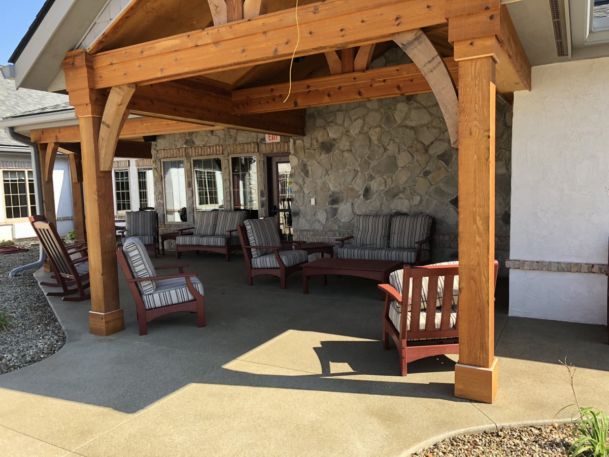A cozy outdoor patio area with wooden beams, featuring cushioned armchairs and a couch against a stone wall.