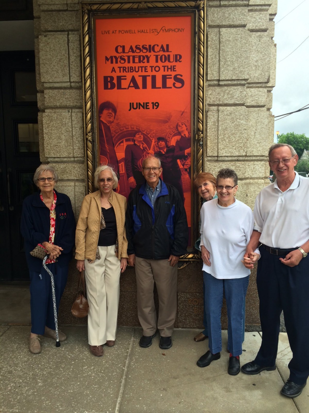 A group of elderly people stand in front of a Beatles poster outside a building.