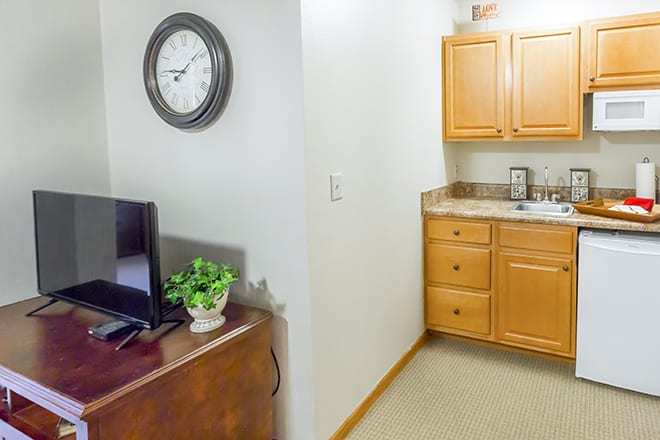 A small kitchen area with wooden cabinets and a TV stand featuring a clock on the wall.