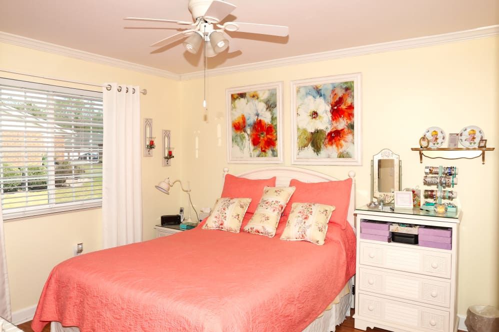 A cozy bedroom with a pink bedspread, floral artwork above the bed, and a ceiling fan.
