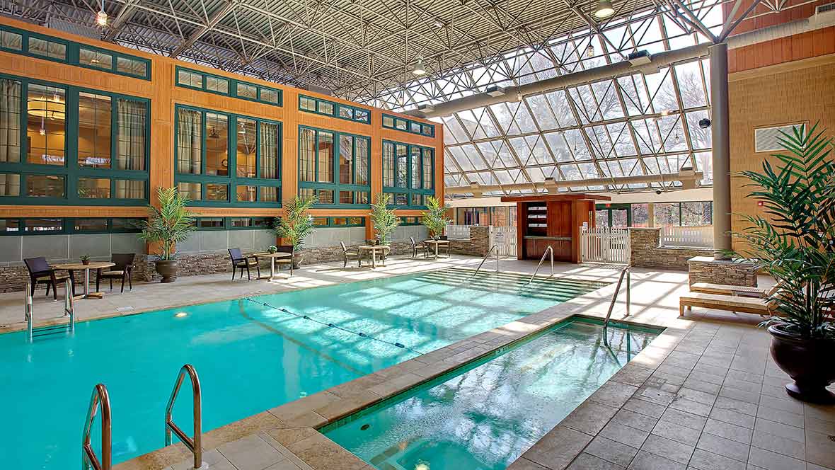A spacious indoor swimming area with a glass ceiling and modern architectural design, featuring a pool and jacuzzi.