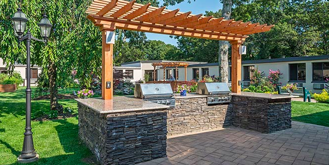 A backyard outdoor kitchen with two grills, a stone countertop, and a wooden pergola.