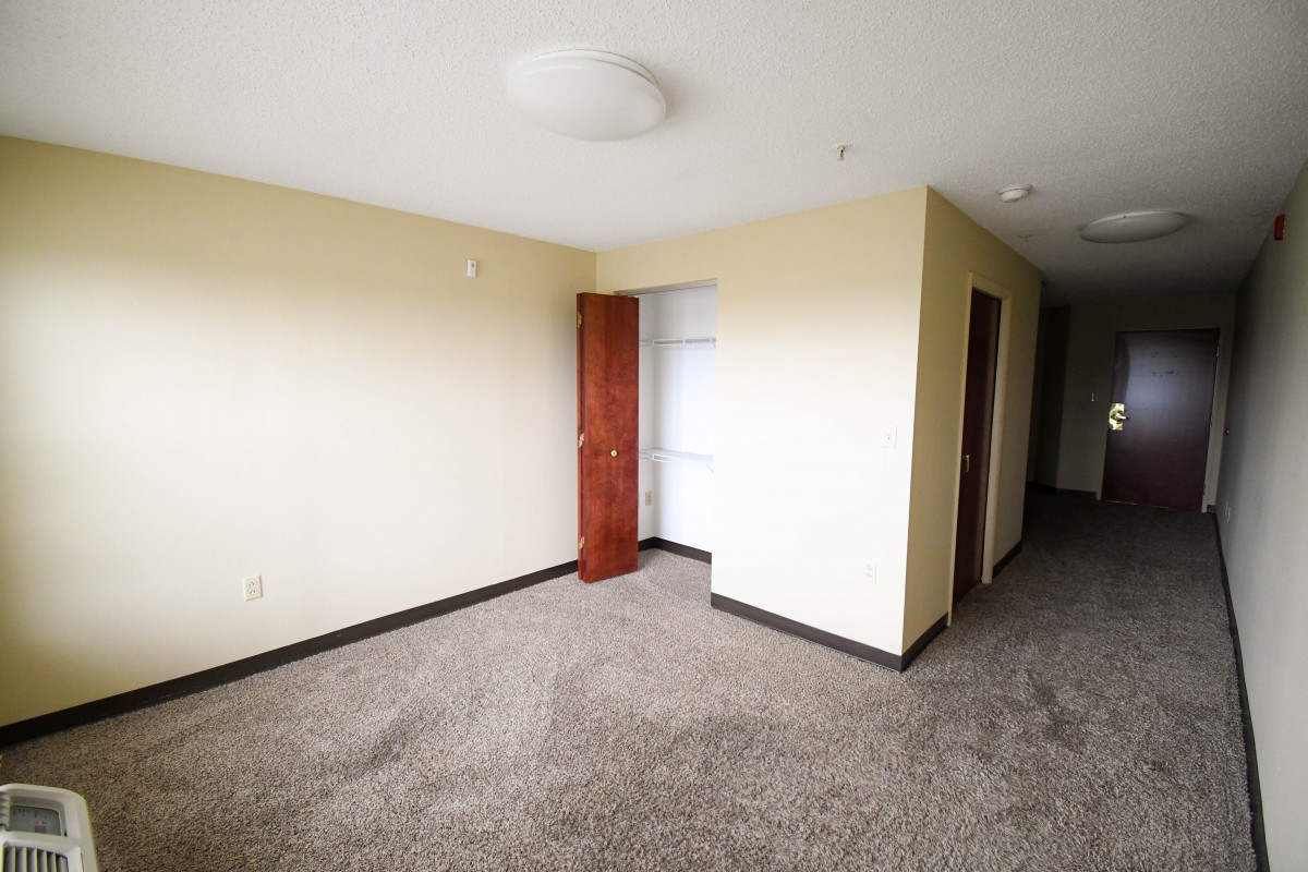 An empty room with carpet flooring and a window, featuring a closed wooden door and a ceiling light fixture.