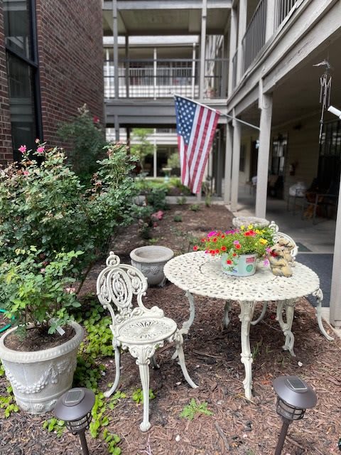 A cozy outdoor patio setup featuring a decorative table and chair, surrounded by potted plants and an American flag.