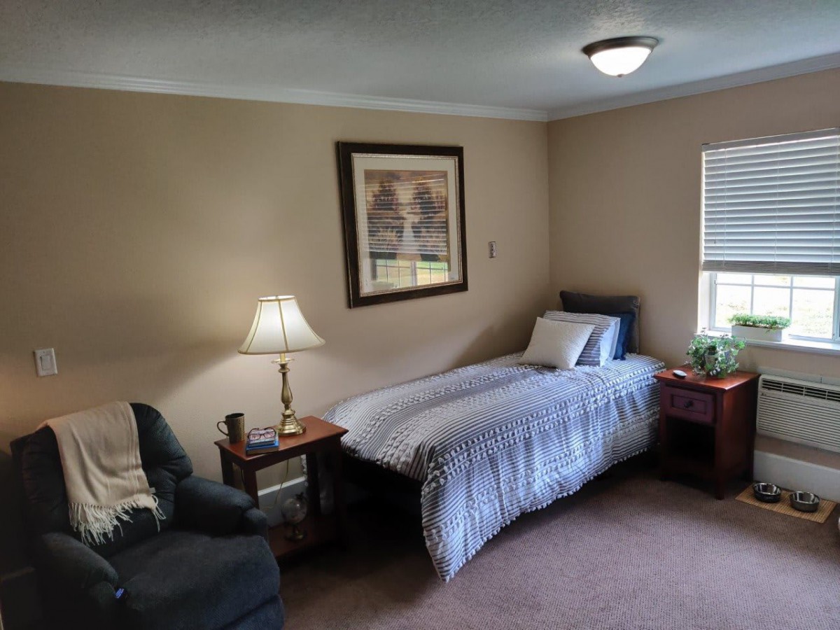 A cozy bedroom featuring a single bed with striped bedding, a lamp on a nightstand, a framed picture on the wall, and a comfortable chair by the window.