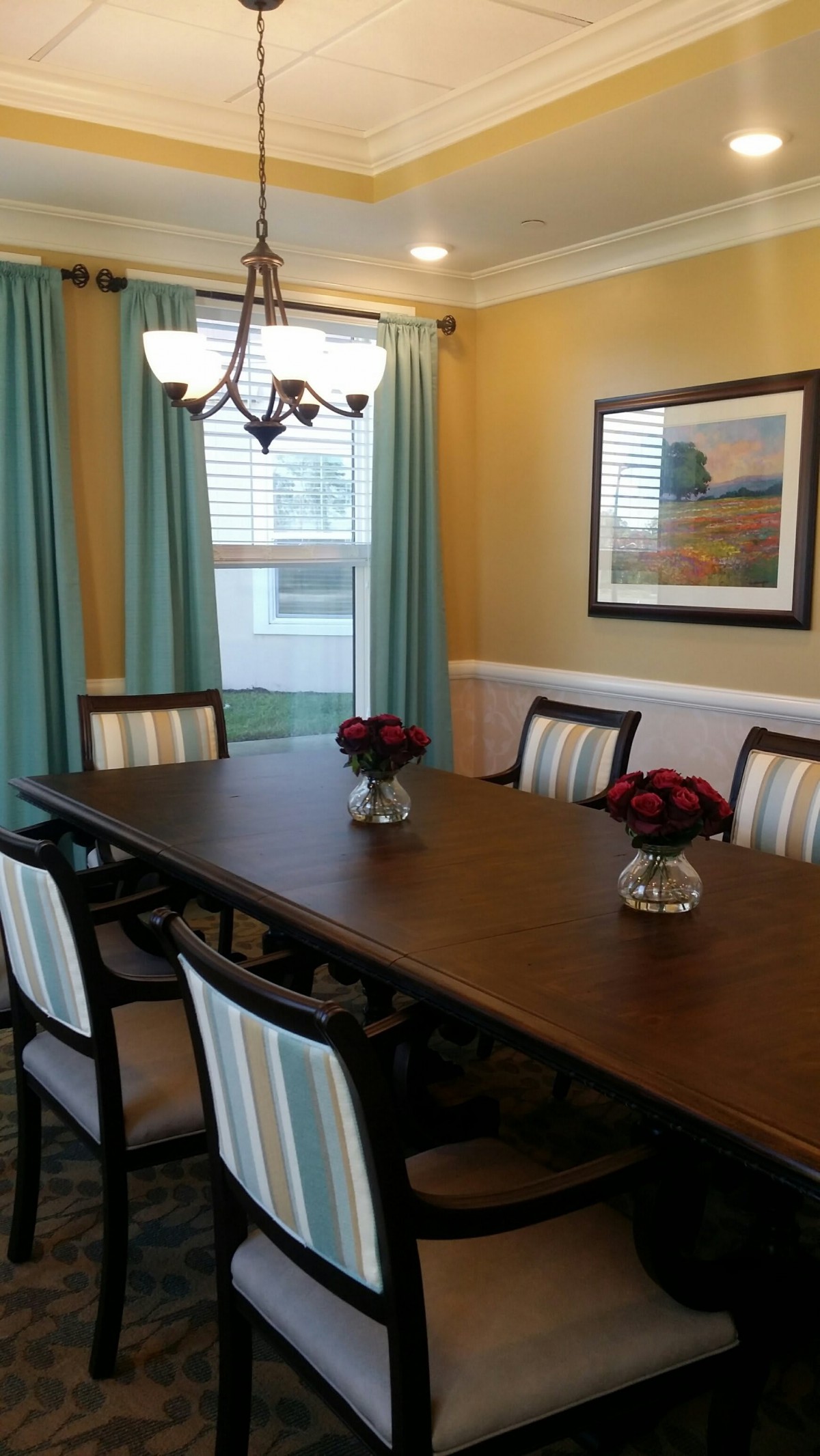 A well-decorated dining room featuring a long wooden table with striped chairs, accented by a chandelier and vases of red flowers.
