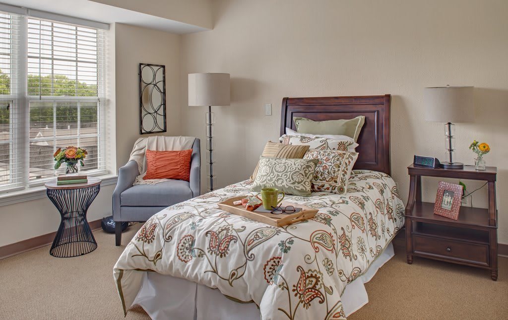 A cozy bedroom with a single bed featuring floral bedding, an armchair, and a lamp by the window.