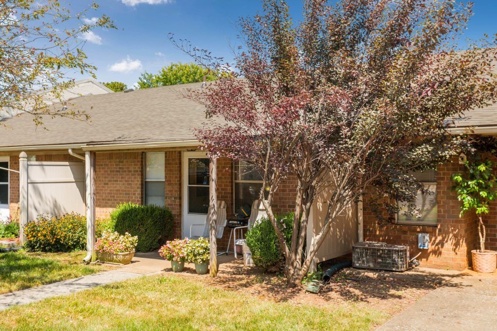 A brick house with a front yard featuring shrubs and trees on a sunny day.