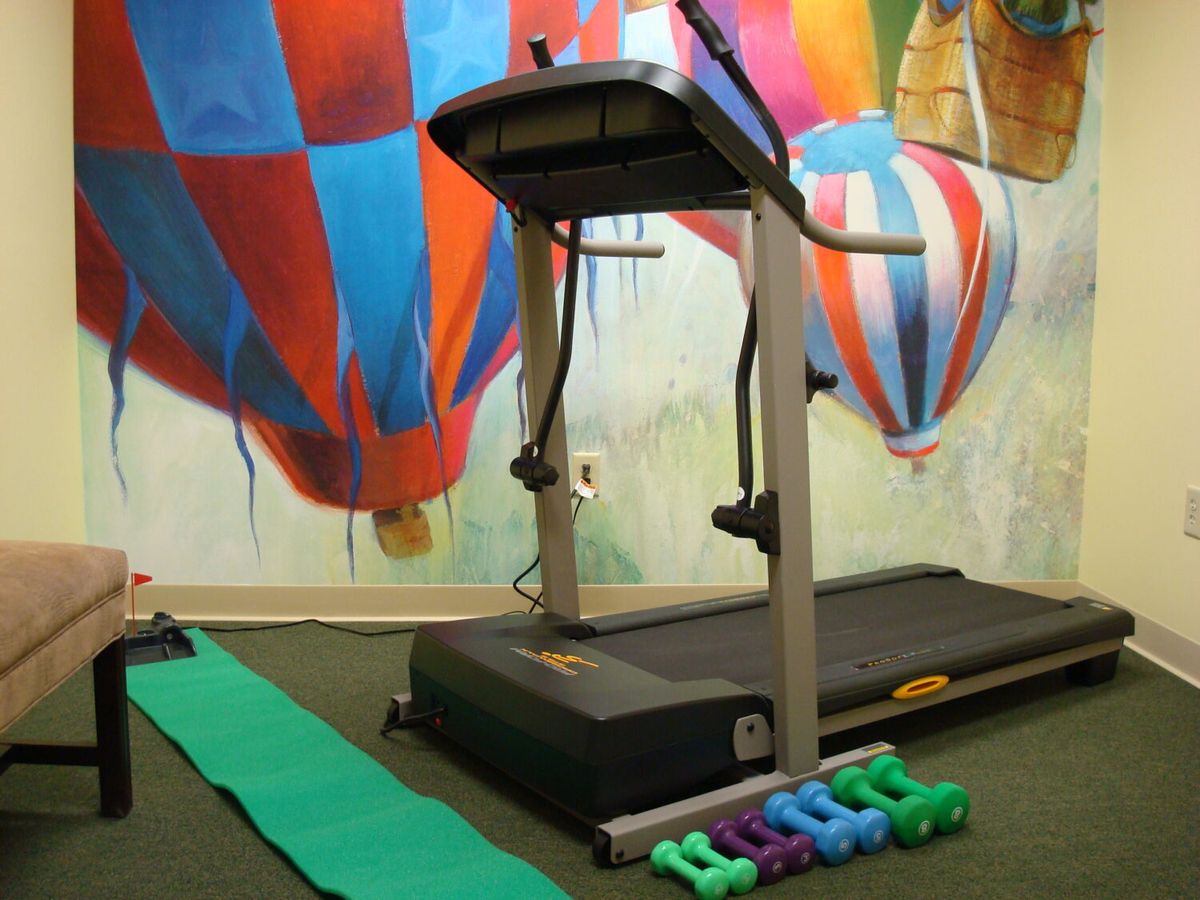 A home gym setup featuring a treadmill, dumbbells, and a yoga mat against a colorful hot air balloon mural.