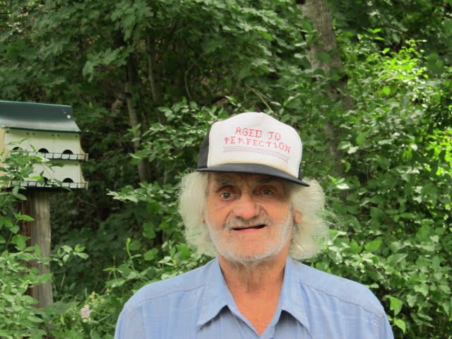 A man wearing a hat stands outdoors with trees in the background.