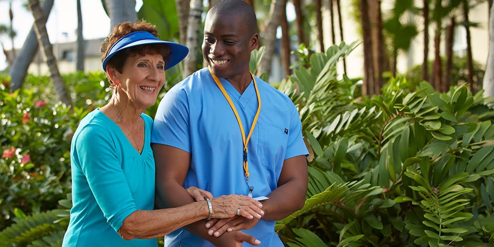 A nurse assists an elderly woman outdoors, surrounded by greenery.