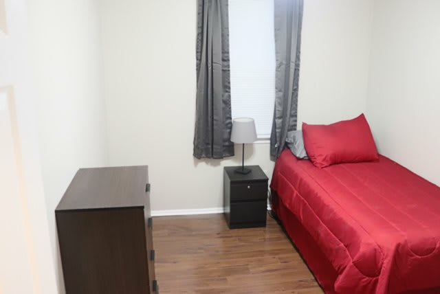 A simple bedroom with a red bedspread, nightstand with a lamp, and a dark wood dresser.