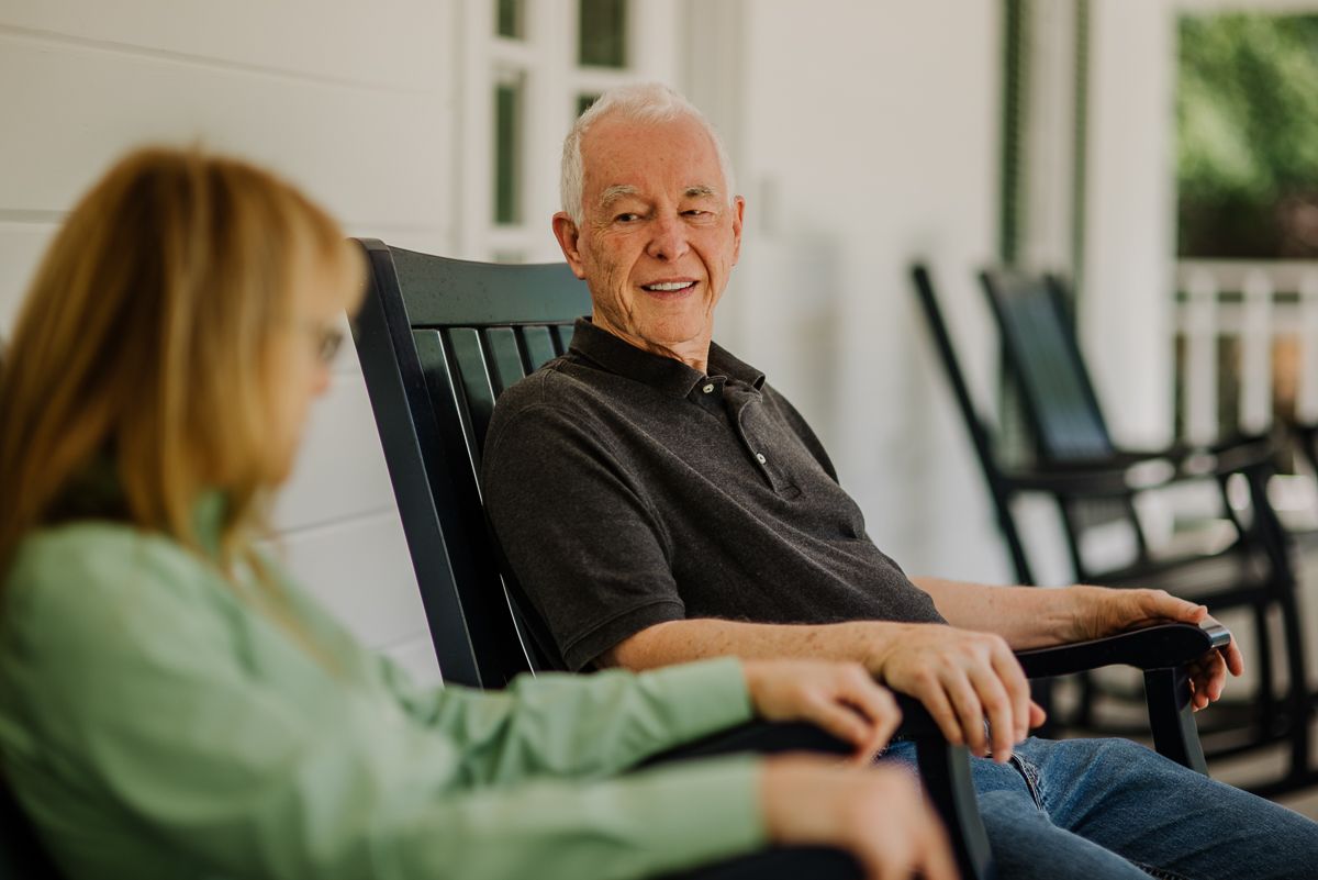 An elderly man and a woman enjoying a conversation on a porch while sitting in rocking chairs.