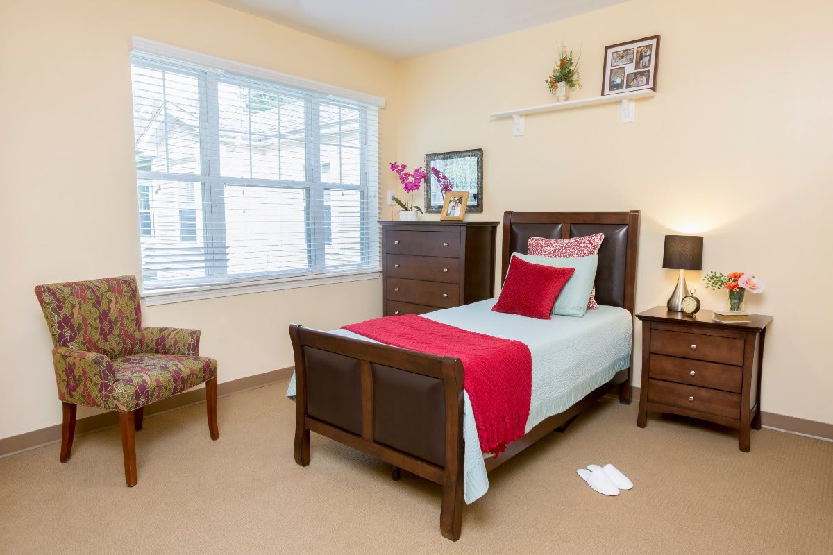 A cozy bedroom with a single bed, adorned with red and white bedding. The room features a window allowing natural light, a wooden dresser, and decorative elements on the walls.