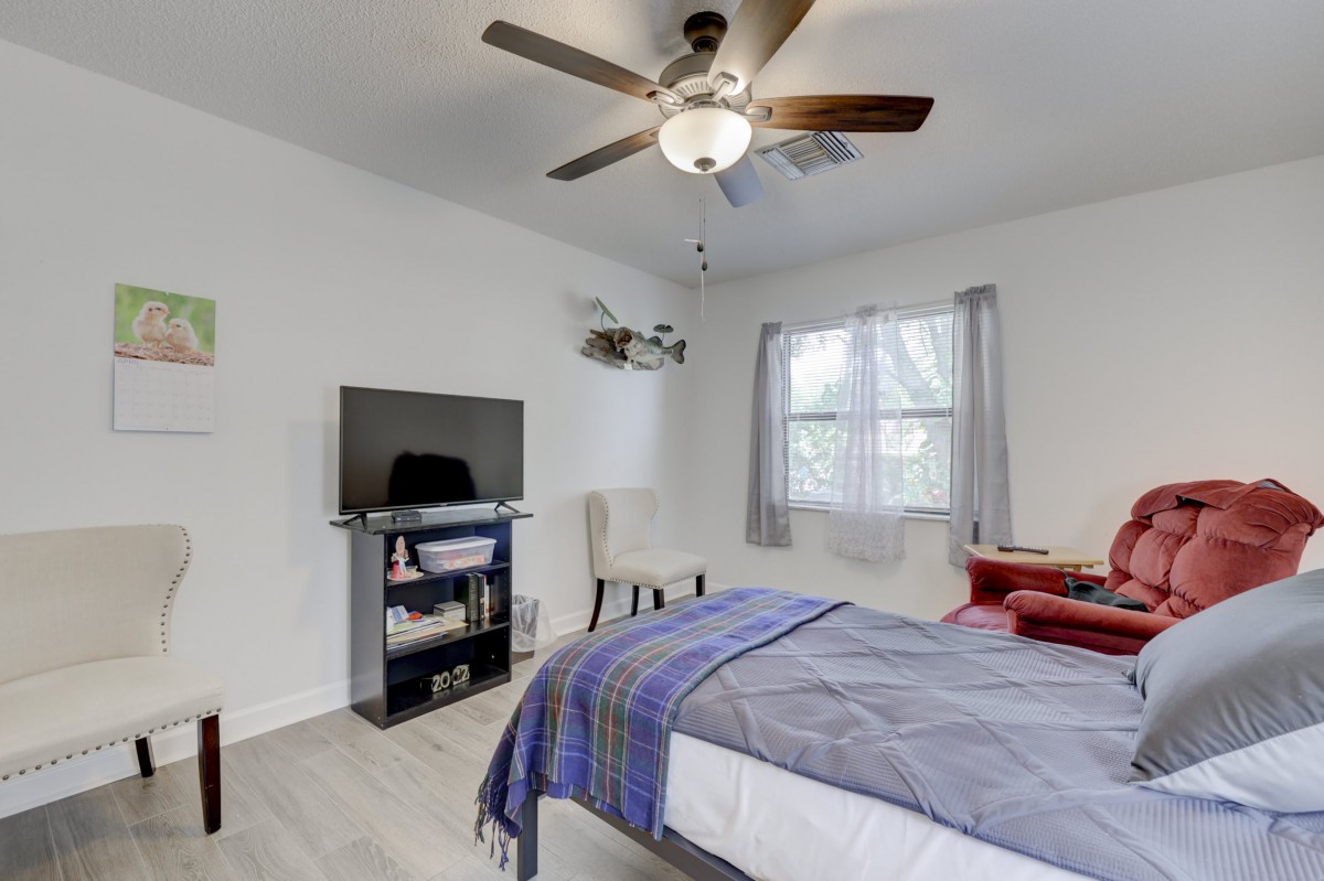 A cozy bedroom featuring a bed with a blanket, a ceiling fan, a television on a stand, and two chairs near a window with curtains.