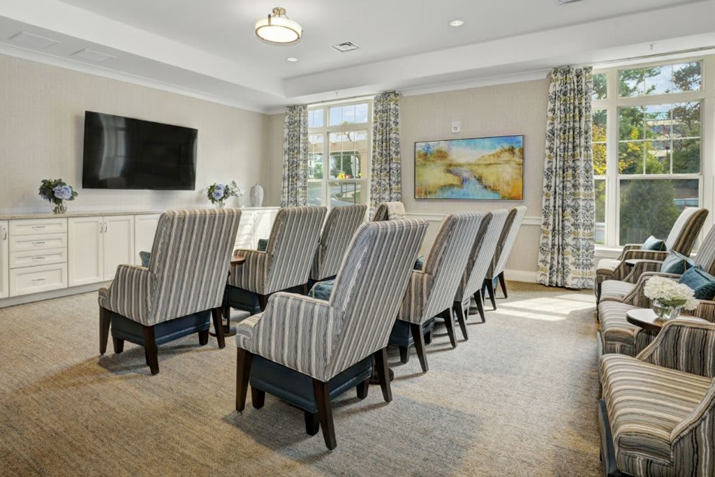 A well-lit conference room with rows of upholstered chairs facing a wall-mounted television, featuring artwork and windows with patterned curtains.