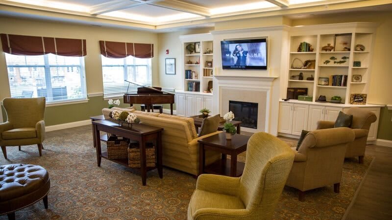 A cozy living room with comfortable seating, a piano, a television above the fireplace, and bookshelves filled with decor.